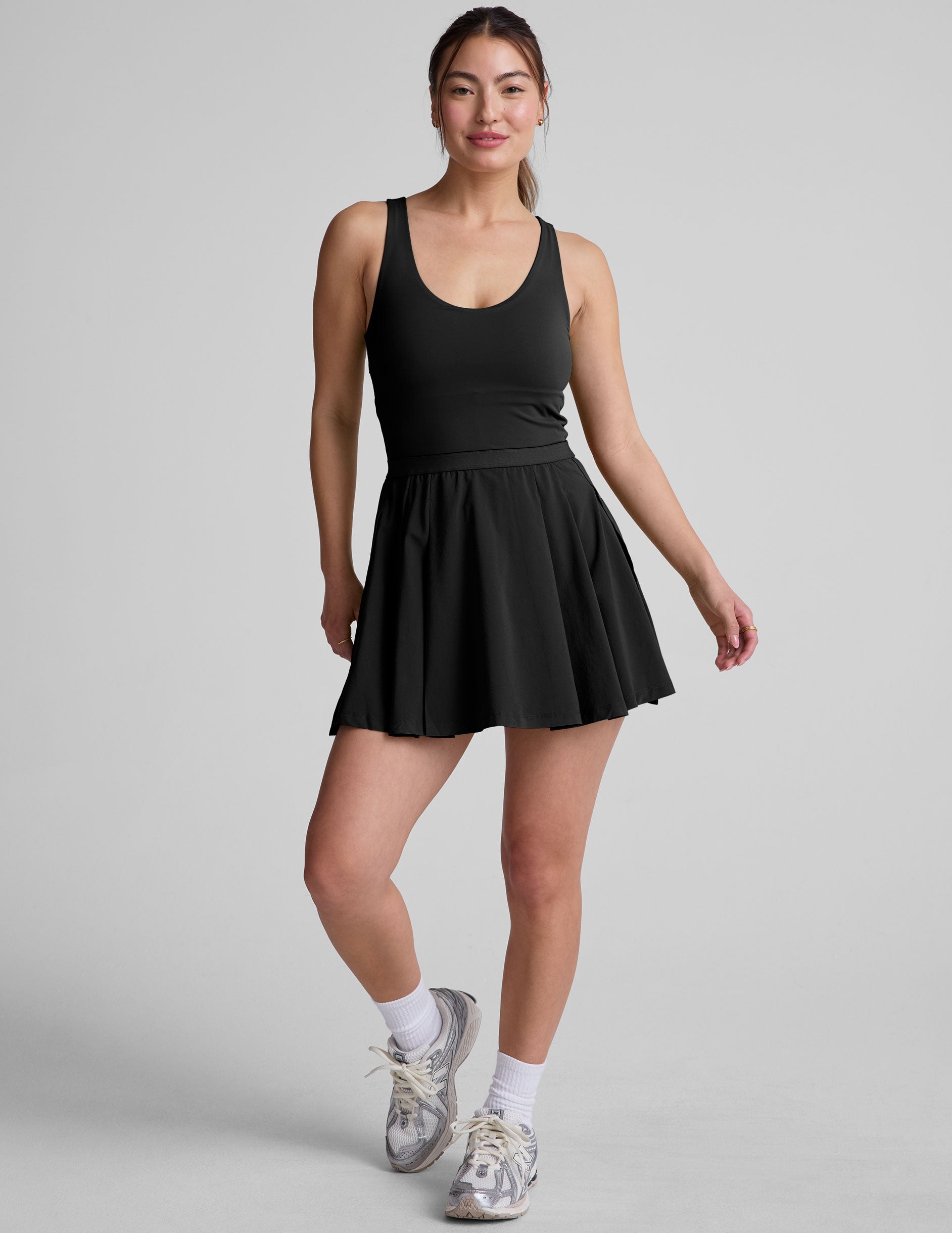black mini dress in our stretch woven fabric with stitch down front pleats, a scoop front neckline, open back detail, bodice in performance stretch fabric, and skirt side vents for easy pocket access.