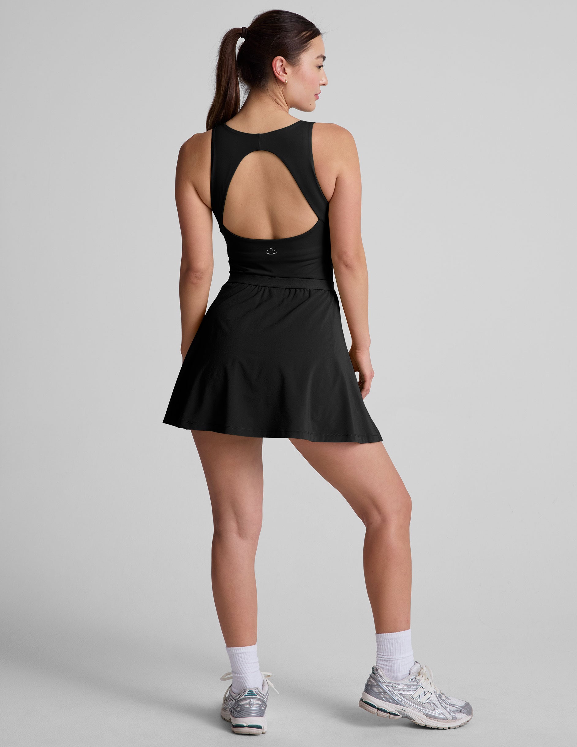 black mini dress in our stretch woven fabric with stitch down front pleats, a scoop front neckline, open back detail, bodice in performance stretch fabric, and skirt side vents for easy pocket access.